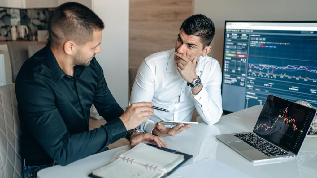 Two cryptocurrency investors analyzing Bitcoin trading charts on laptop discussing investment strategy during market crash